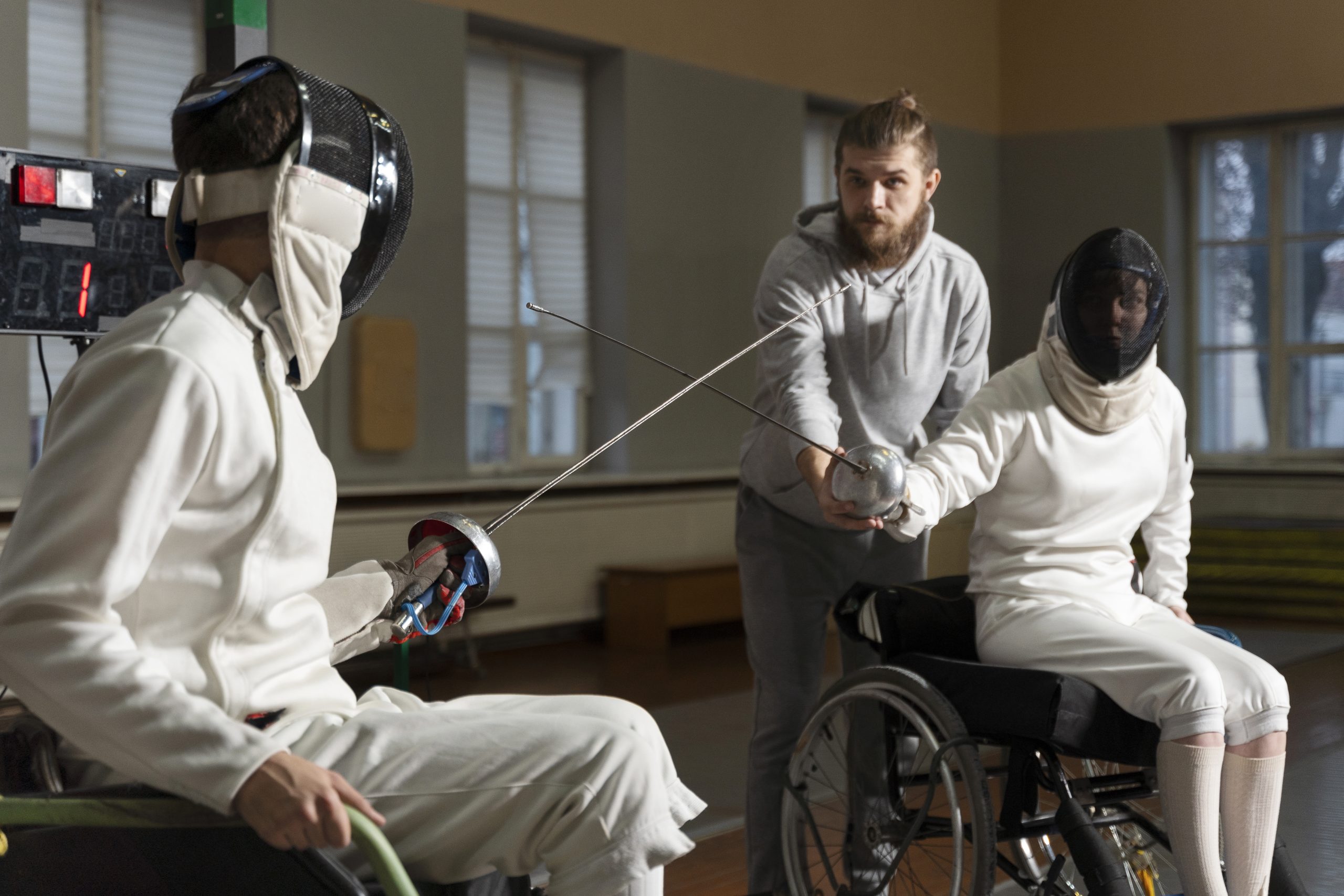 Entraînement d’escrime en fauteuil roulant avec deux athlètes équipés et un coach corrigeant leurs positions.